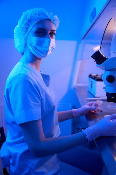 彼女の手で液体溶液で満たされたバイアルで顕微鏡の前に座っている穏やかな女性科学者 — ストック写真