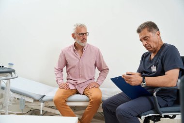 Ultrason odasında muayene olan gri saçlı bir adam. Doktor hastaların şikayetlerini kaydediyor.