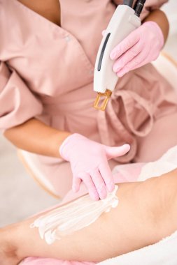 Kozmetikçi, modern bir cihaz kullanarak, lazer tüy çıkarma prosedüründen önce hastaların ellerini koruyucu kremle yağlıyor.