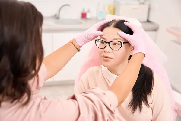 Female cosmetologist examines the forehead of a patient, a woman wearing pink clothes