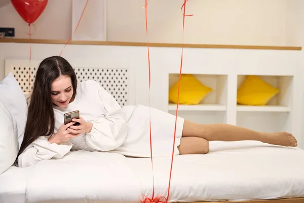 Concentrated female in white robe using cell phone on cozy bed with red balloons in hotel room