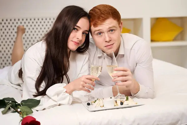 若い女性と男性は ホテルの部屋でベッドに横たわっている間 カメラを見てシャンパンのグラスを保持しています — ストック写真