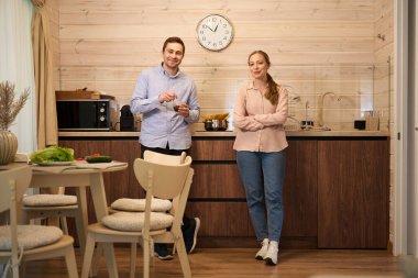 Sıcak kahve içen bir adamın portresi ve mutfakta duran sağlıklı yemek pişiren bir kadın.