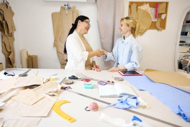 Terzi, moda tasarımcısını iş yerinde karşılar, kadınlar kesim masasında durur.