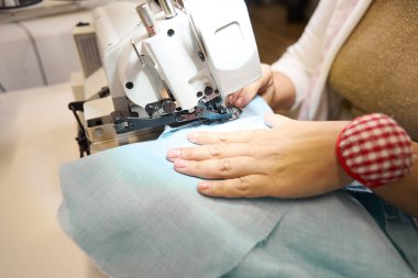 Terzi mavi kumaşlı bir dikiş makinesinde çalışıyor, işyeri iyi aydınlatılmış.