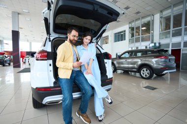 Beyaz kadın ve erkek, araba galerisinde araba bagajına yaslanırken gazeteyi inceliyor.