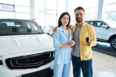 Elinde anahtar arabasıyla yakışıklı bir adam ve şık giyinmiş güzel bir kadın araba galerisinde kameraya bakıyor.