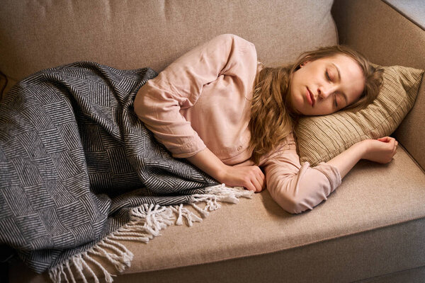 Pretty young woman laying on couch covered with blanket while sleeping peacefully during day