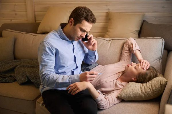 Unhealthy tired lady with cold touching forehead lying under plaid on sofa while her boyfriend checking thermometer