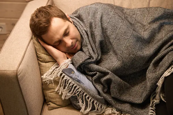 Tired male laying on comfortable couch covered with blanket while sleeping peacefully on soft pillow