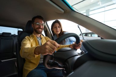 Yakışıklı bir adam arabanın içinde oturuyor ve elinde direksiyon tutarken kız arkadaşı da onu araba merkezinde izliyor.