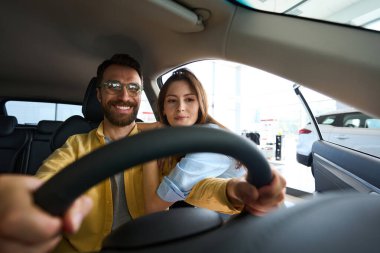 Gülümseyen adam arabanın içinde oturuyor ve elinde direksiyon tutarken kız arkadaşı onunla araba merkezinde flört ediyor.