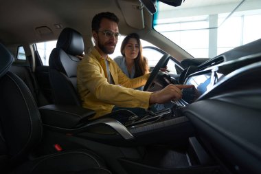 Yakışıklı bir adam arabanın içinde oturuyor ve elinde direksiyon tutarken kız arkadaşı da onu araba galerisinde izliyor.