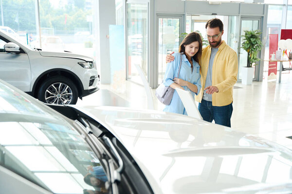 Молодая женщина и мужчина любуются белым автомобилем, стоя в ярком автосалоне