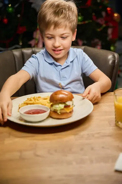 Little boy looking at big burger on his plate while sitting at table in restaurant on Christmas dinner