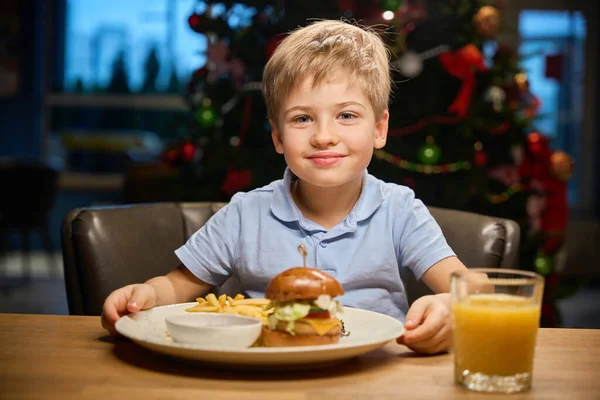 Cute little boy eating burger while sitting in restaurant during winter holidays