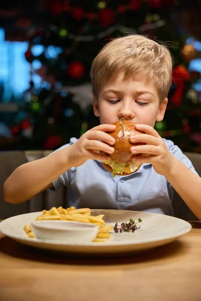 Little boy biting big burger enjoying it taste while celebrating Christmas in restaurant