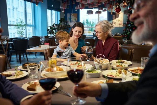 Happy senior woman sitting at table with little boy enjoying Thanksgiving dinner with her family in restaurant