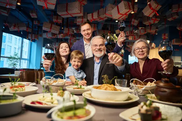 Overjoyed family holding glasses with wine smiling happily celebrating Christmas together in restaurant