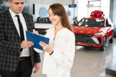 Otomobil galerisinde araba satıcısı olan genç ve mutlu bir kadın yeni otomobil satın alma belgelerini imzalıyor.