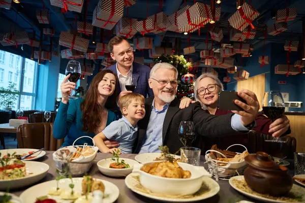 お祝いのレストランでクリスマスを祝っている間 セルフィーを取ったり スマートフォンでビデオ通話をしたりする楽しい家族 — ストック写真