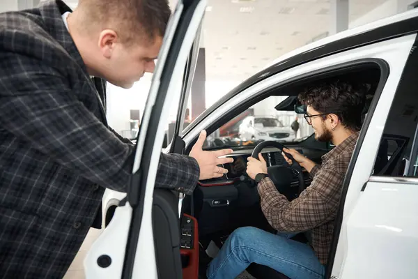 Young man buying new automobile in showroom talking with manager about car models
