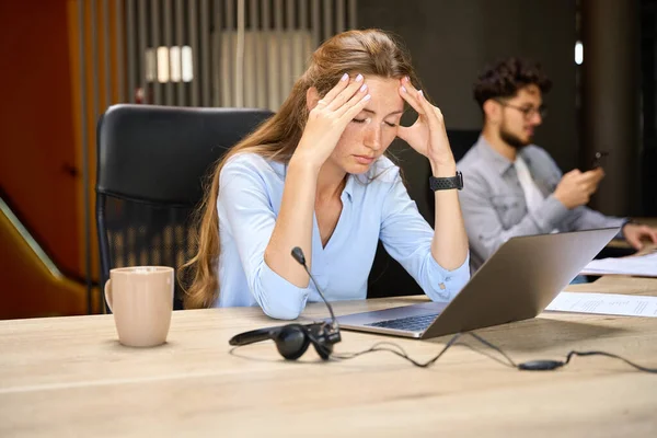 年轻疲惫的 闭眼的高加索女商人在同事办公室里与模糊的男性同事一起在笔记本电脑前工作时感到头疼 工作狂和工作倦怠 — 图库照片