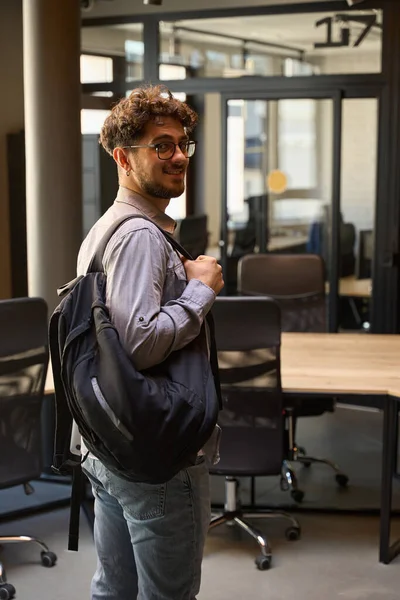 Young smiling caucasian businessman with backpack and wearing glasses standing and looking at camera in empty coworking office. Concept of modern business lifestyle