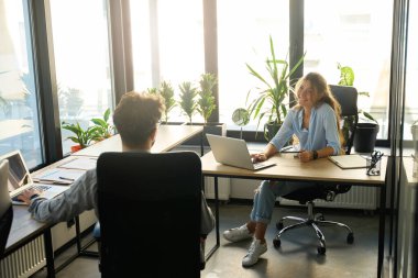 Back of businessman and smiling caucasian businesswoman looking at each other while working on laptops at desks in sunny office. Concept of teamwork