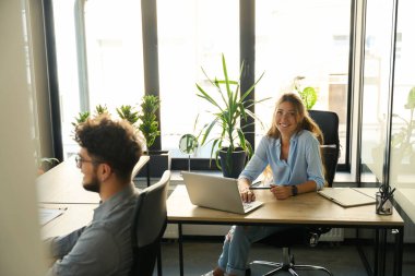 Young smiling european businesswoman looking at camera while working on laptop with her male colleague in sunny office. Concept of modern business lifestyle