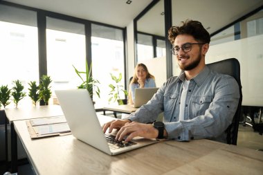 Young smiling european businessman wearing glasses working on laptop with her blurred female colleague on backgrounds in sunny office. Concept of modern business lifestyle