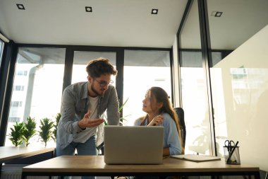 Businesswoman looking at male colleague while he showing something on laptop to her during work in sunny office. Concept of teamwork