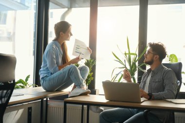 Side view of smiling businesswoman showing business graph to focused male colleague in sunny office. Concept of teamwork