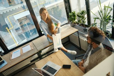 High angle view of businesswoman with business graph and her male colleague looking at each other in sunny office. Concept of teamwork