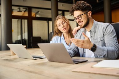 Young smiling caucasian businessman showing something on laptop to female colleague during work at desk in coworking office. Concept of teamwork