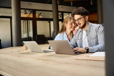 Young smiling businesswoman holding hand on shoulder of shocked caucasian male colleague watching on laptop during work at desk in coworking office. Concept of teamwork