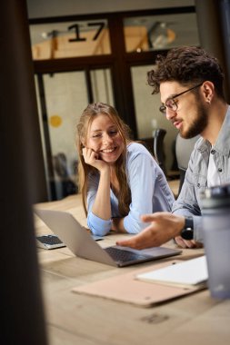 Young caucasian businessman showing something on laptop to smiling female colleague at desk in coworking office. Concept of teamwork