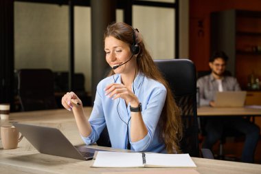 Young smiling female caucasian call center operator watching on laptop during work at desk with her blurred male colleague on background in coworking office. Concept of modern business lifestyle