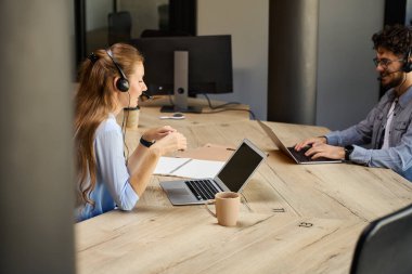 Side view of young smiling caucasian call center operators using laptops during work at desk in coworking office. Concept of teamwork