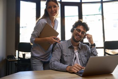 Young smiling businesswoman looking at camera while standing near laughing male colleague wearing headset using laptop in office. Concept of teamwork