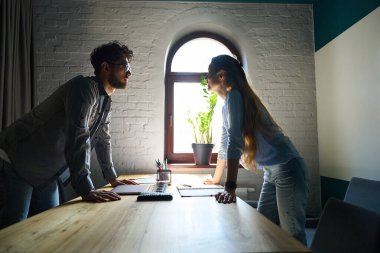 Side view of young serious european businessman and businesswoman standing and looking at each other while working at desk in coworking office. Concept of teamwork