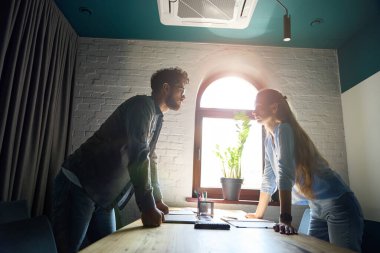 Side view of young serious caucasian businessman and businesswoman standing and looking at each other while working at desk in sunny coworking office. Concept of teamwork