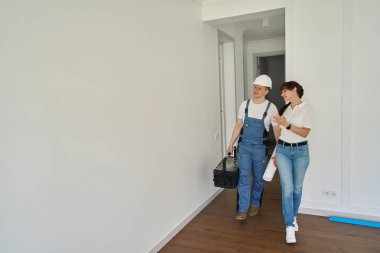 Smiling woman showing new modern comfortable townhouse for concentrated repairman with blueprint and toolbox before repair