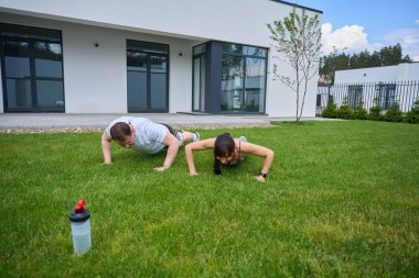 Adult caucasian couple doing push ups on green grass in yard near modern townhouse in warm summer day. Family relationship and spending time together. Healthy lifestyle