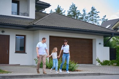 Joyful caucasian mother, father and daughter holding hands and going from new modern townhouse outdoors in summer day. Family future planning, relationship and spending time together