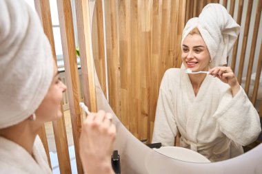 Woman brushes her teeth in front of a mirror, she is in a fluffy bathrobe