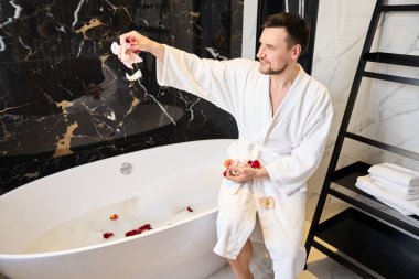 Smiling male preparing a bath with rose petals, man in a fluffy bathrobe
