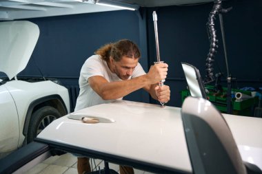 Man uses a special tool to level out body dents, a man fixes a white car