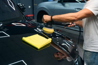 Mechanic in car repair shop polishes the hood of a black car, he uses a grinder and a soft cloth