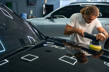 Long-haired young man polishes the body of a black car, he uses a special device